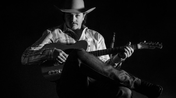 Black & white image of a man sat holding a guitar wearing a cowboy hat, shirt, jeans and boots